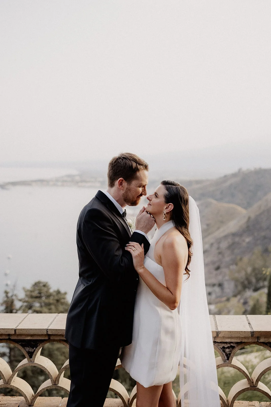 A bride and groom share an intimate moment on a balcony with scenic hills and a body of water in the background.