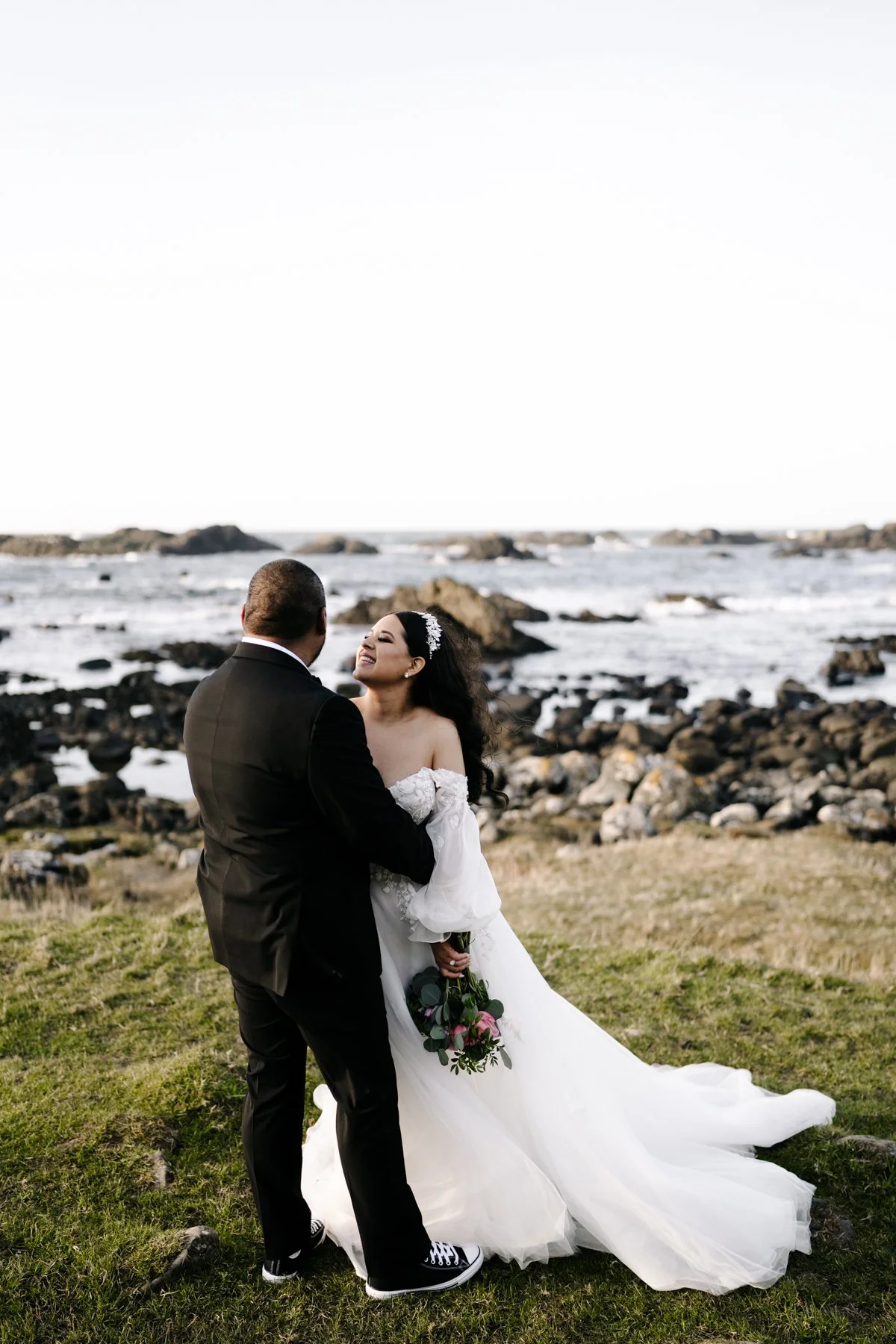 A bride and groom standing outdoors near the ocean, with rocks and waves in the background, sharing a joyful moment in wedding attire.