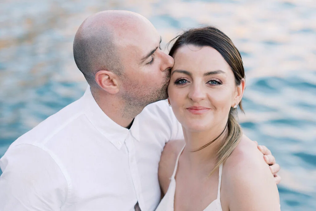 A man is kissing a woman on the cheek by a body of water, both smiling softly.