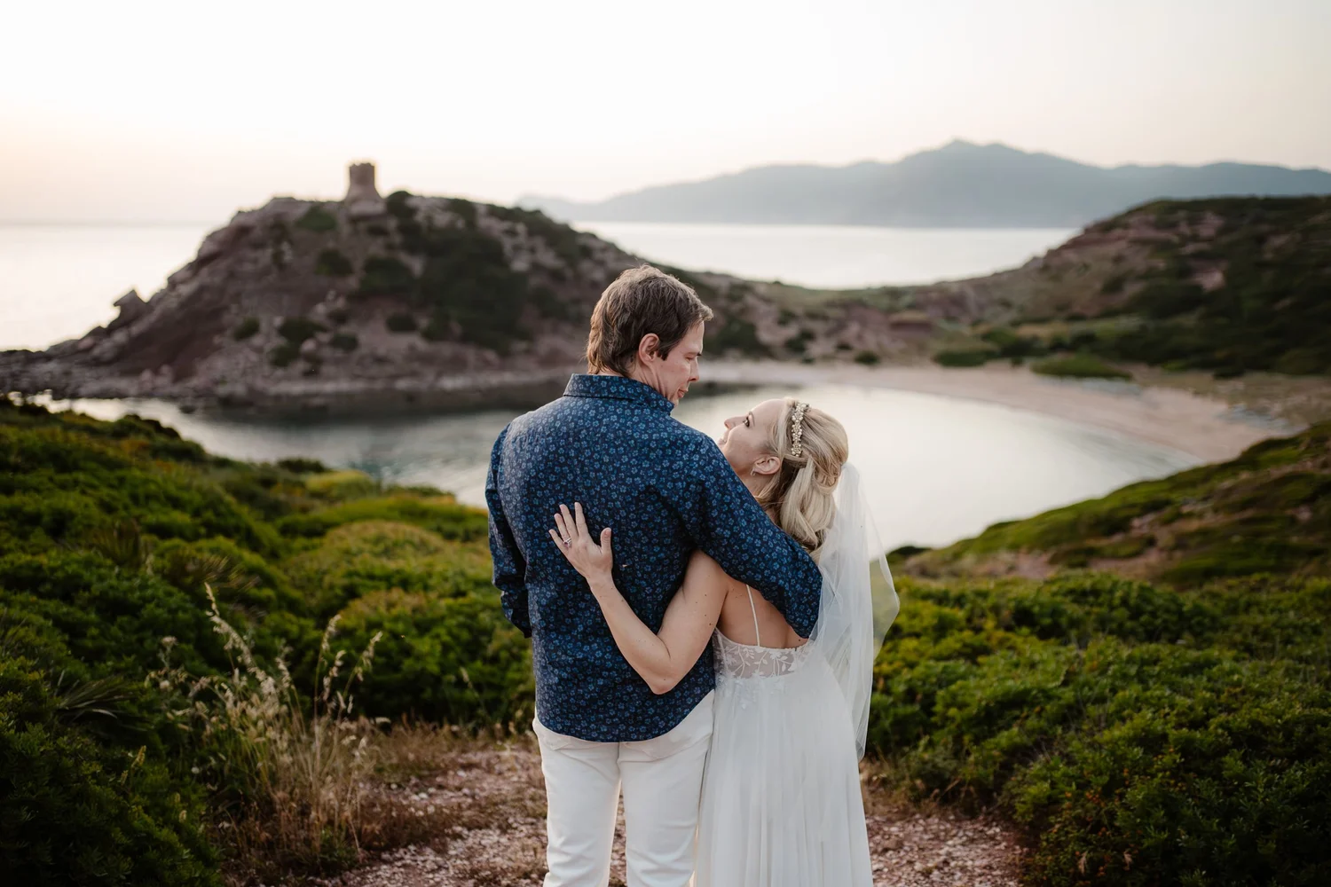 A man and woman dressed in wedding attire embracing outdoors near water and green hills, with mountains in the background.