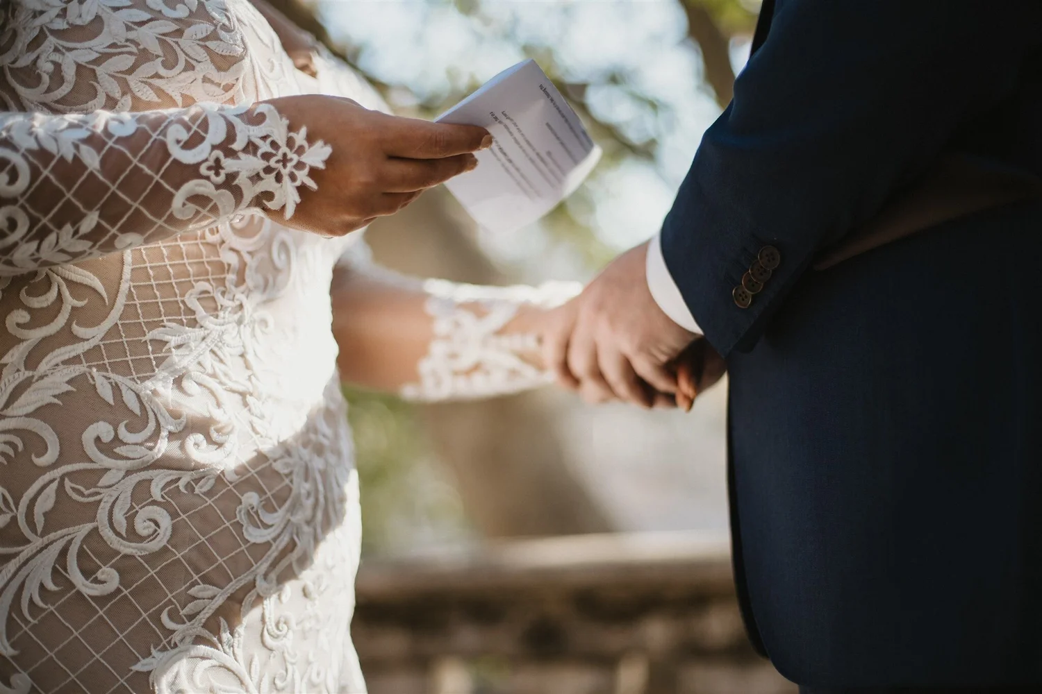 A bride and groom holding hands during a wedding ceremony outdoors. The bride is wearing a white lace dress with floral embroidery, and the groom is dressed in a navy blue suit with a white shirt.
