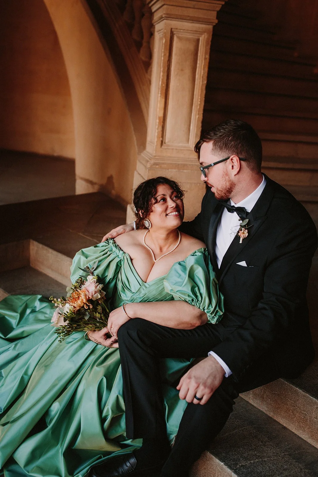 A woman in a green dress sitting on steps beside a man in a tuxedo, both gazing at each other affectionately in a warmly lit, wood-paneled setting. The woman holds a bouquet of flowers and wears jewelry, including a pearl necklace.