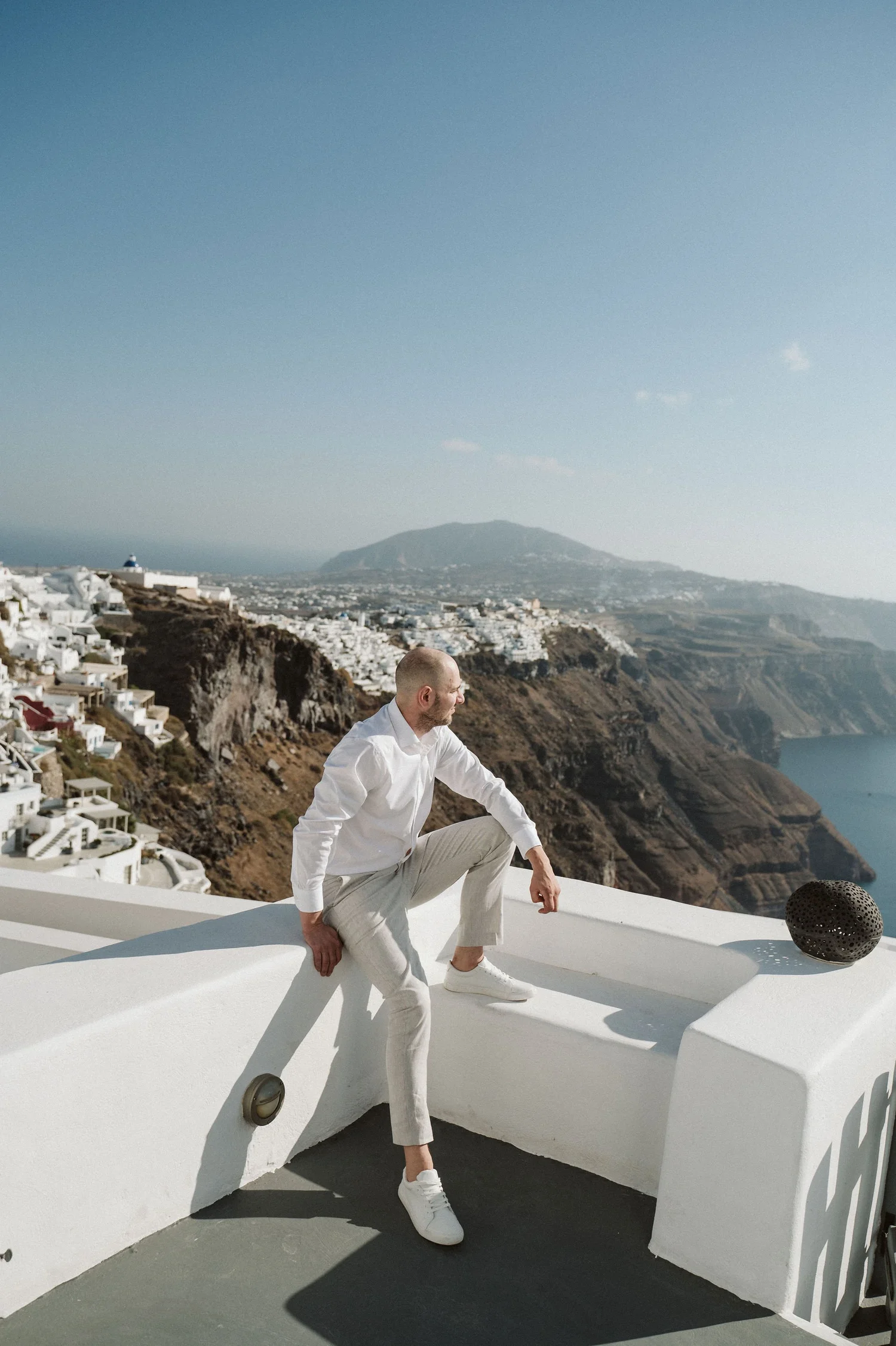 A man in white clothing and sneakers sitting and relaxing on a white ledge overlooking a coastal town with white buildings built on hillside, and a mountain in the background.