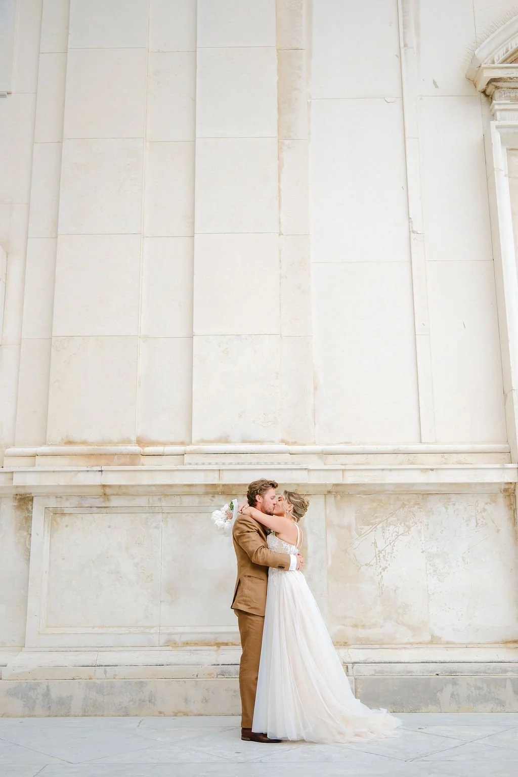 A bride and groom embracing and kissing in front of a large marble wall at a wedding, with the bride holding a bouquet of white flowers.