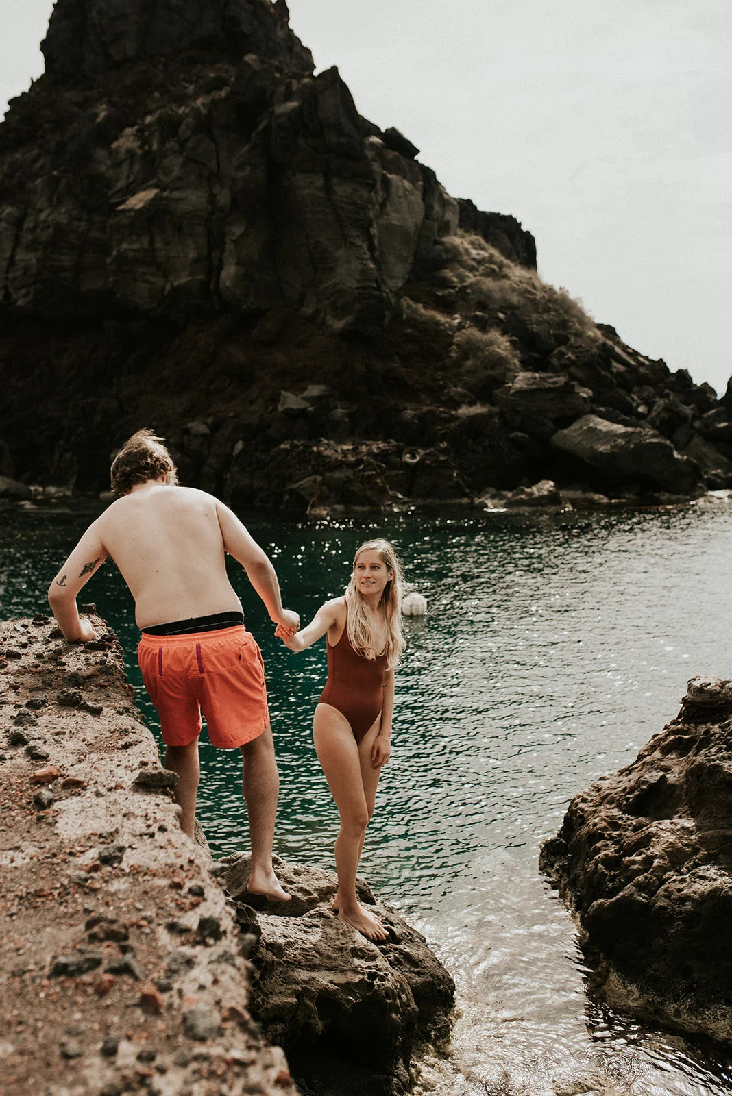 A man and a woman by a rocky shoreline, holding hands, with a body of water and large rocky cliffs in the background.