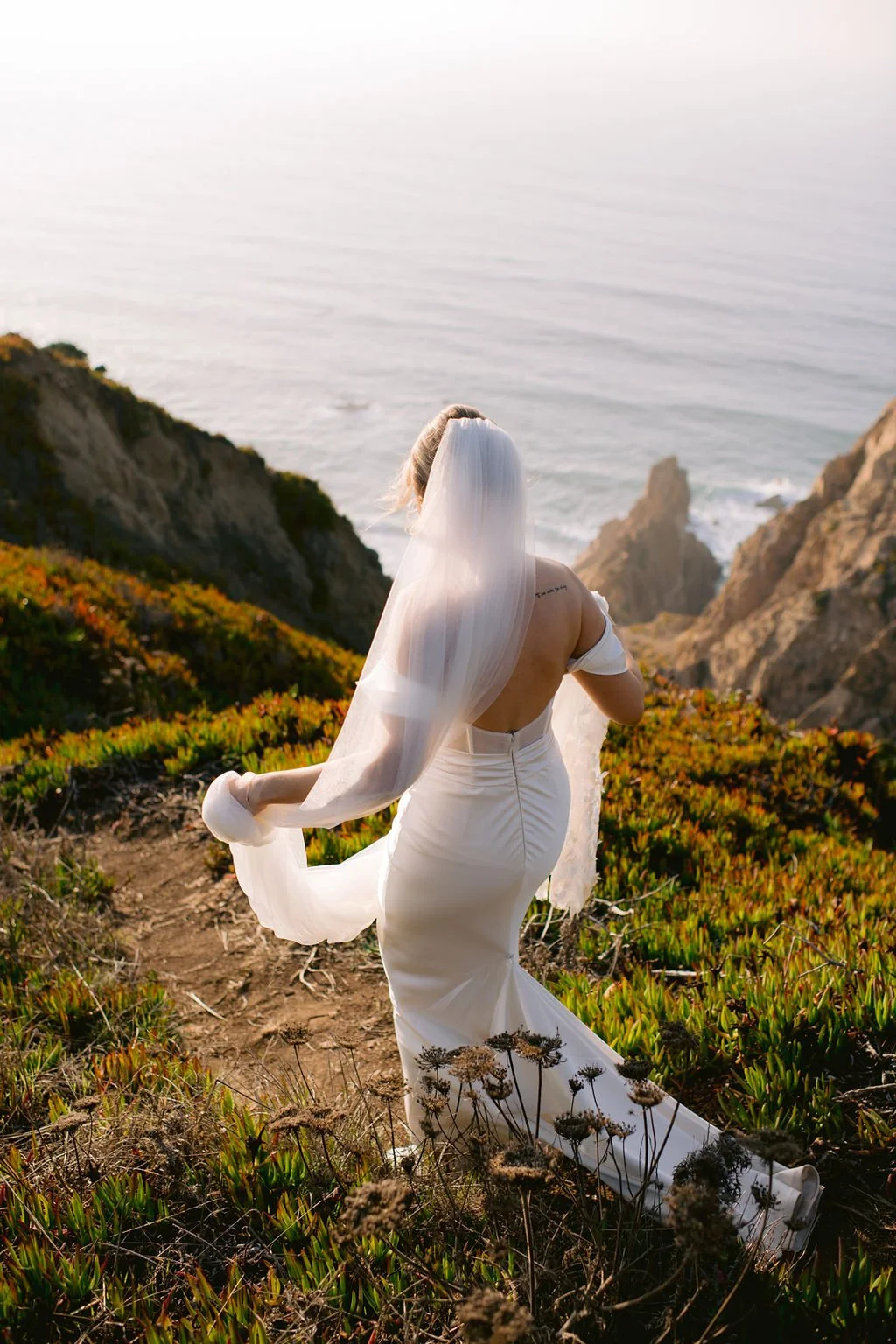 A bride in a white wedding gown with a long train and veil standing on a rocky hillside near the ocean, captured from behind during sunset.