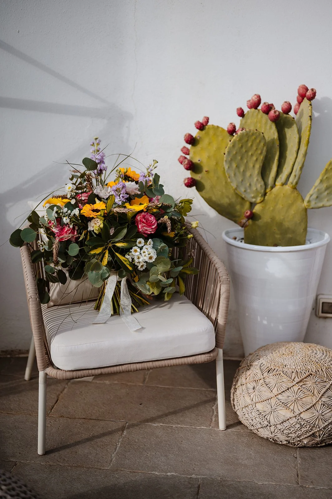 A bouquet of colorful flowers resting on a chair next to a large potted cactus with pink fruit balls, and a decorative round cushion on the floor.