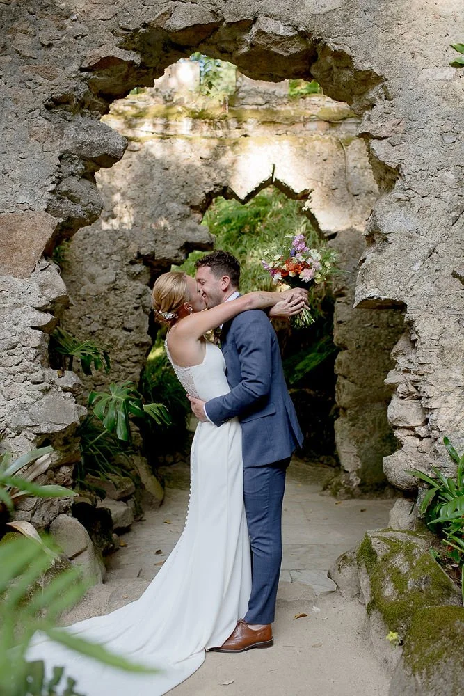 A bride and groom sharing a kiss under an archway made of stone, with greenery and plants around them.