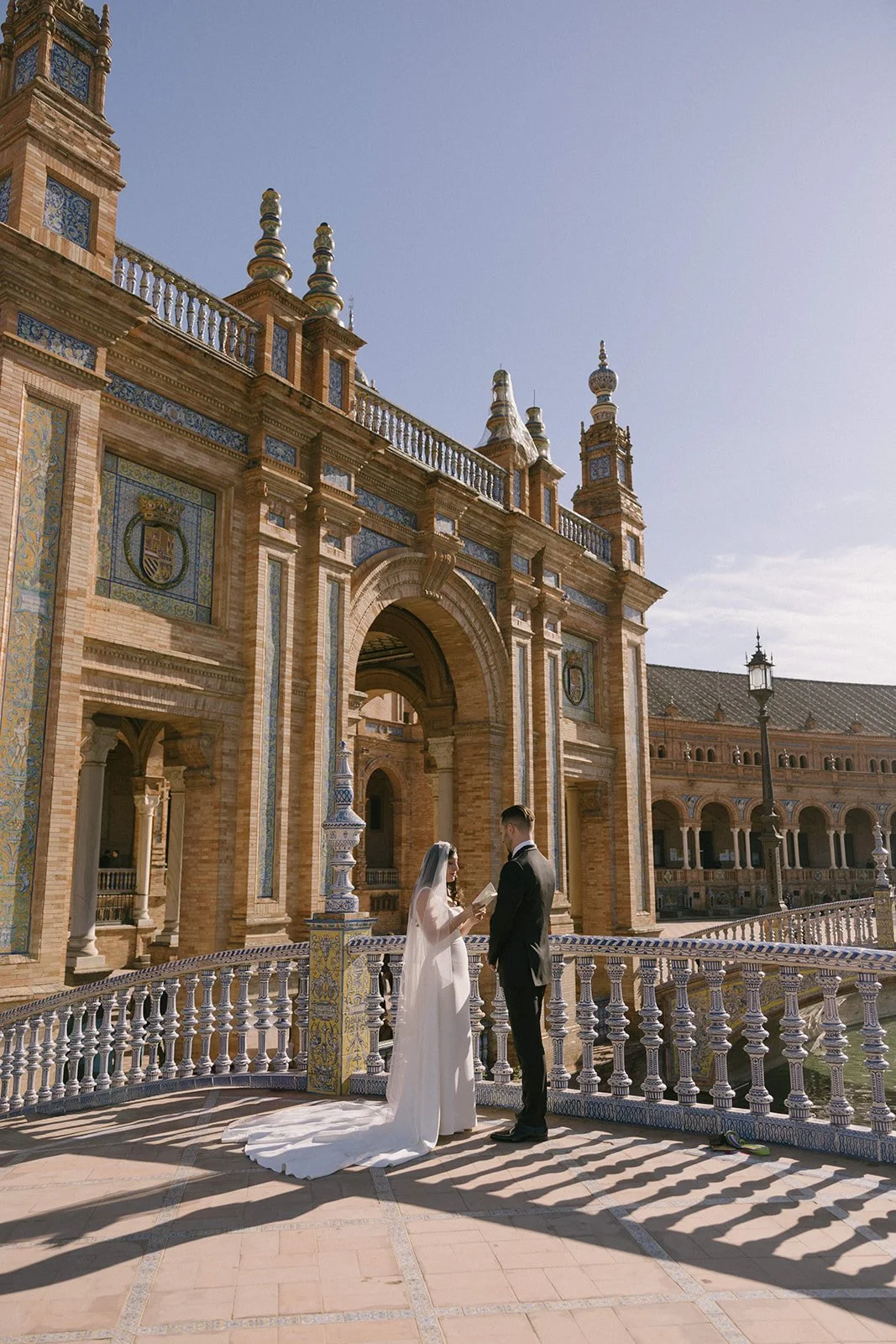 A bride and groom stand on a decorative balcony in front of a historic, ornate building with intricate brickwork and mosaic details, during a wedding ceremony or photoshoot.