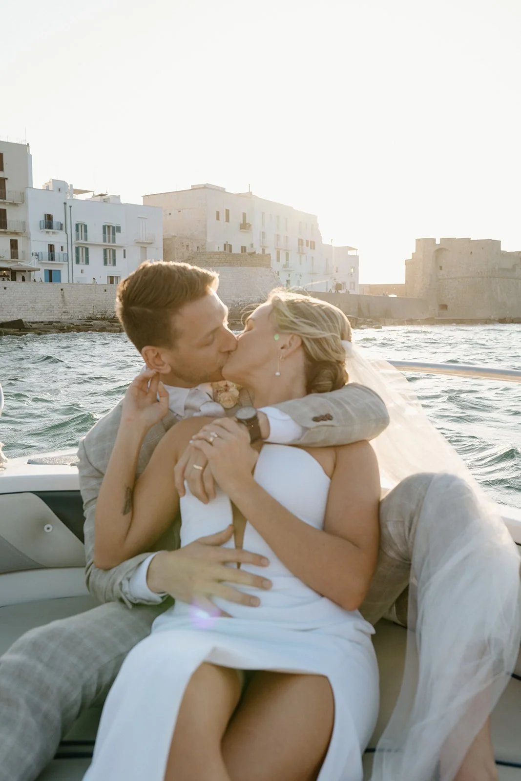 A couple sharing a kiss on a boat during sunset, with a waterfront cityscape of white buildings in the background.