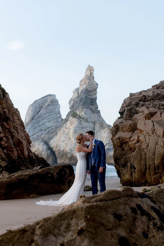Bride and groom sharing a kiss on a beach surrounded by rocks, with large rock formations in the background during sunset.