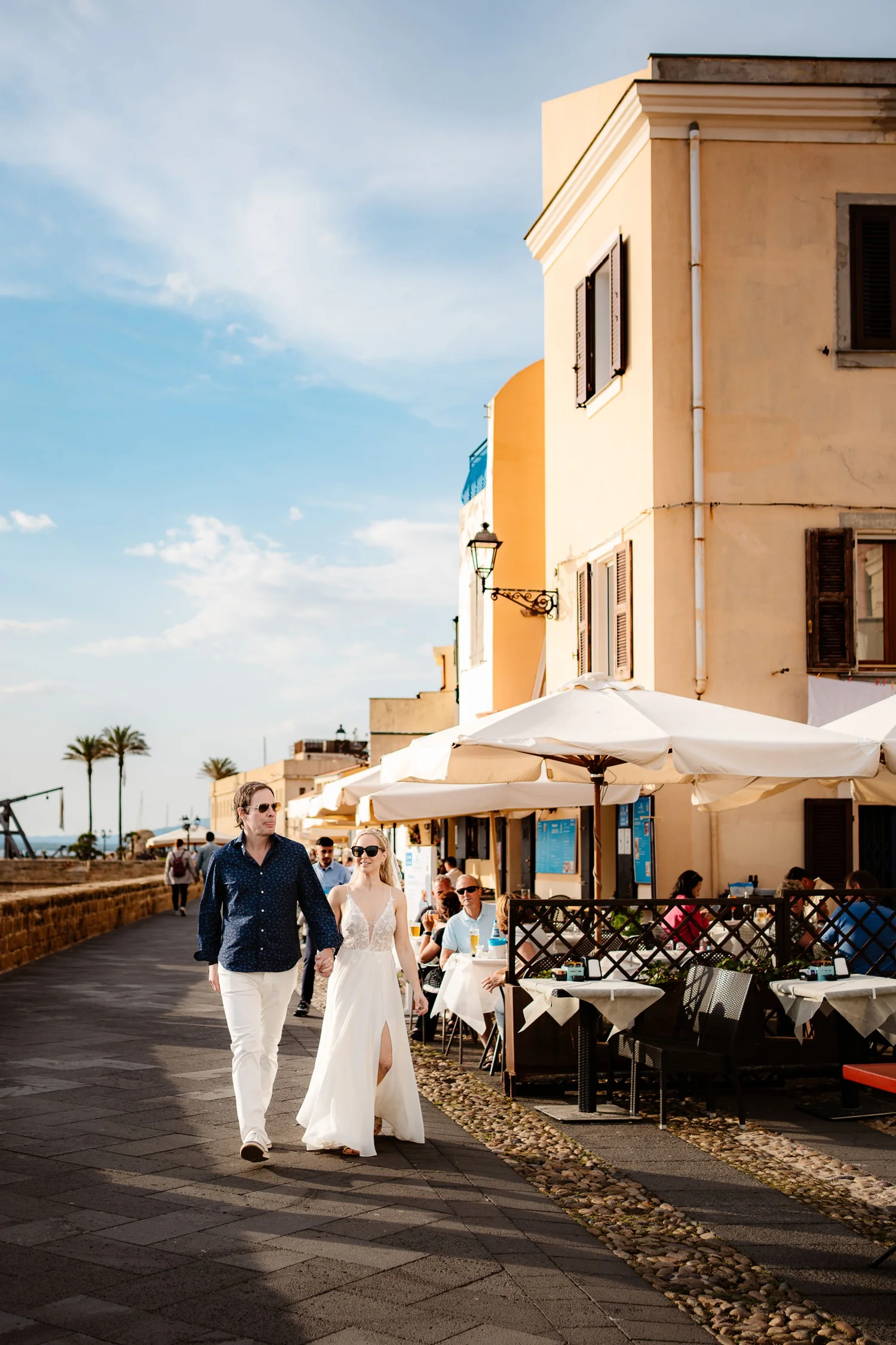 People walking along a seaside promenade with outdoor dining at cafes, yellow and orange buildings, blue sky with clouds, and palm trees in the background.