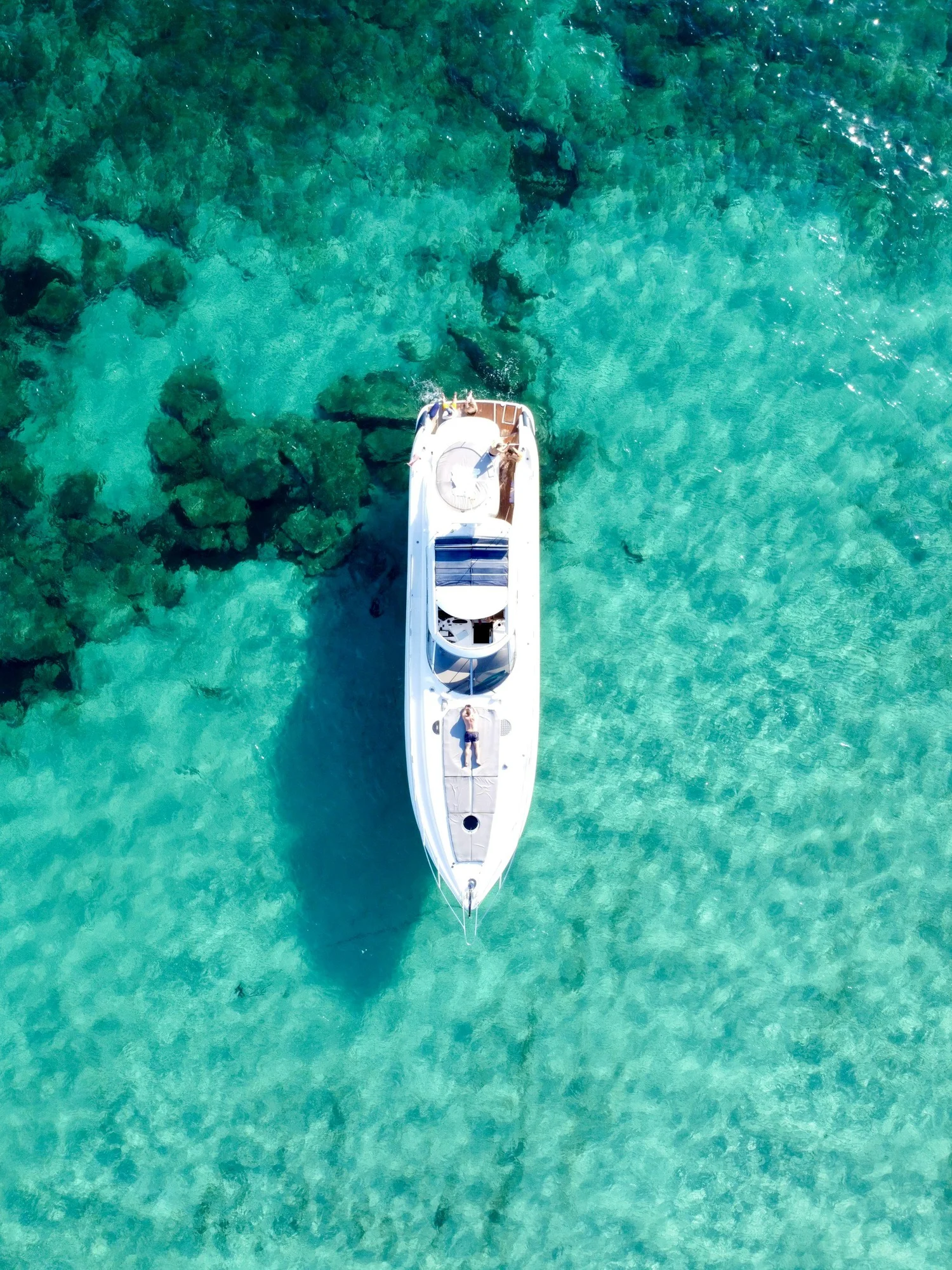 An aerial view of a white yacht floating on clear turquoise water with coral reefs visible beneath the surface.