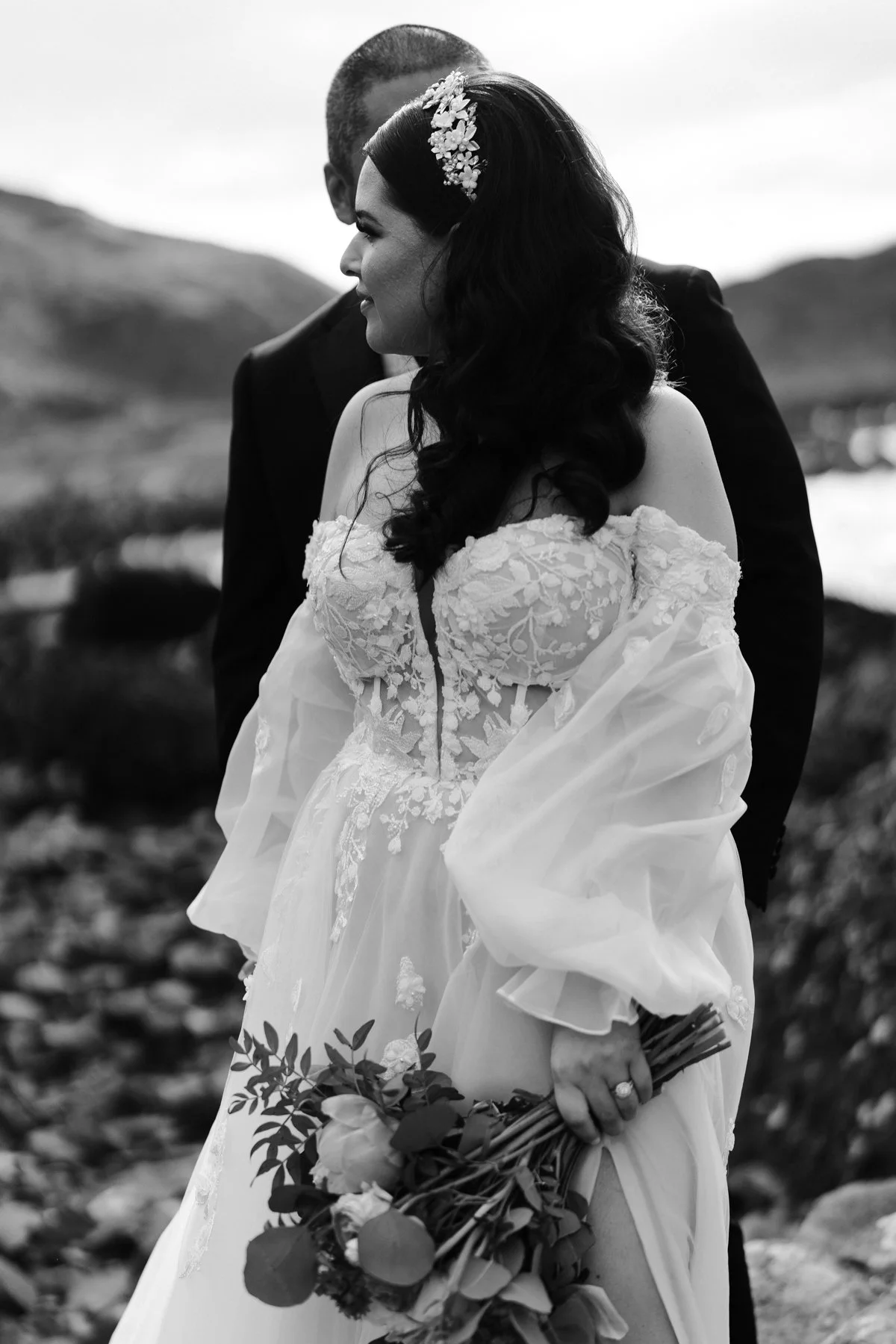 A black-and-white photo of a bride in a lace wedding dress and floral headpiece holding a bouquet, with a groom dressed in a suit, outdoors with mountains and water in the background.