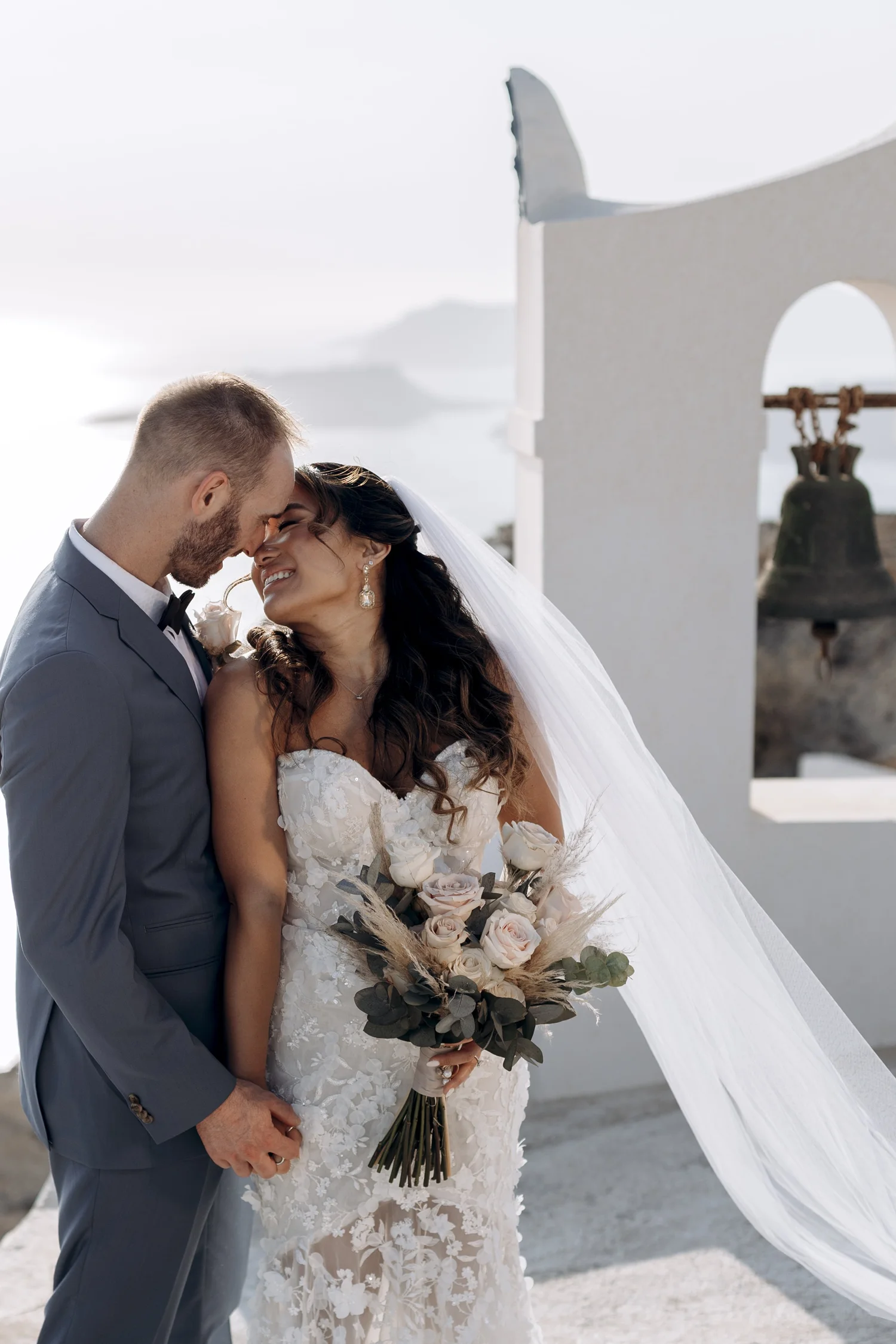 A newlywed couple, a man in a gray suit and a woman in a white lace wedding dress, sharing a joyful moment with their foreheads touching outdoors, with a white wall and bell in the background.