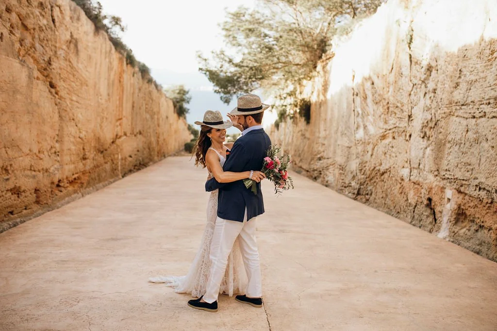 A bride and groom embrace on a dirt road between tall rocky cliffs, with the bride holding a bouquet of flowers.
