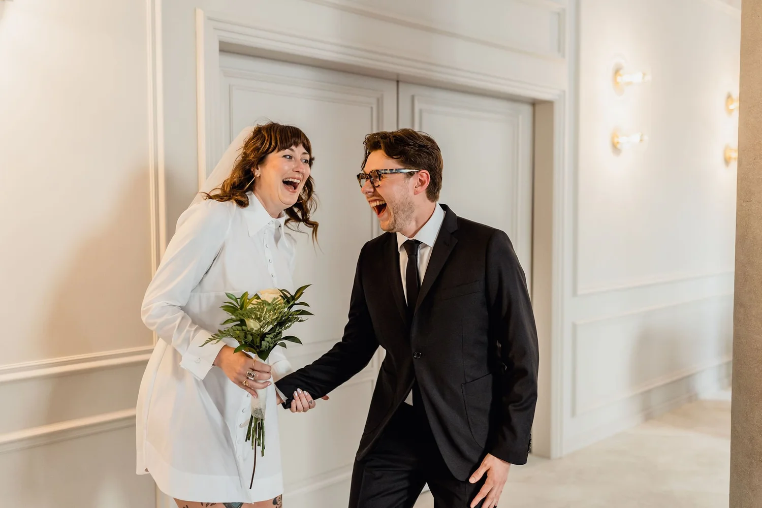 A woman in a white blazer and a man in a black suit share a joyful moment, holding hands and smiling in a well-lit room with white paneled walls. The woman holds a bouquet of white flowers and greenery.