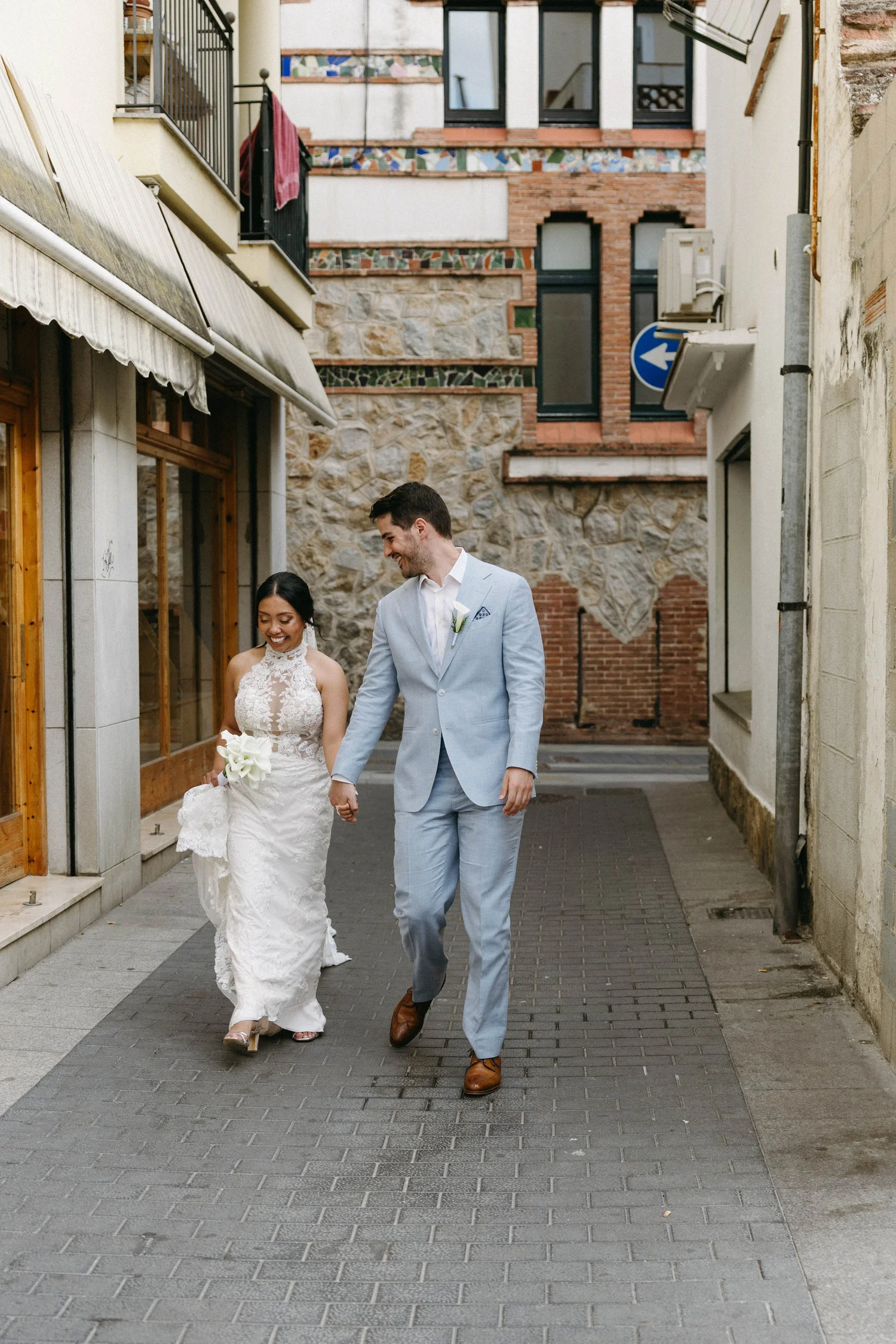 A newlywed couple walking hand in hand down a narrow street, smiling and looking at each other. The bride is wearing a white lace wedding dress and holding a bouquet, while the groom is dressed in a light blue suit with a white shirt. They are surrou