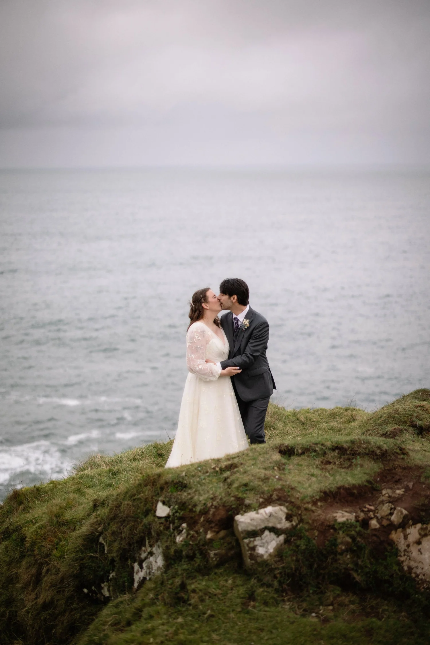 A couple in wedding attire sharing a kiss on a grassy cliff overlooking the ocean on a cloudy day.