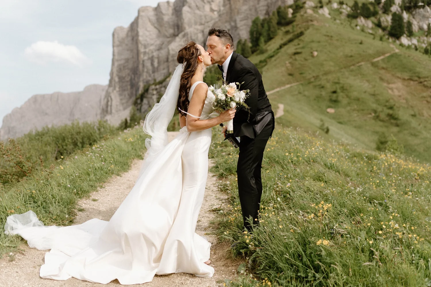 A bride and groom share a kiss outdoors on a trail with mountains and green hills in the background. The bride wears a white wedding gown and holds a bouquet, while the groom wears a black tuxedo.