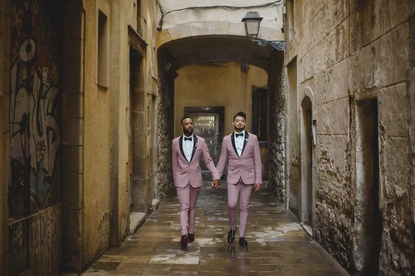 Two men in matching pink tuxedo suits holding hands walk through a narrow alleyway with graffiti on the left wall and weathered stone on the right, under an archway.