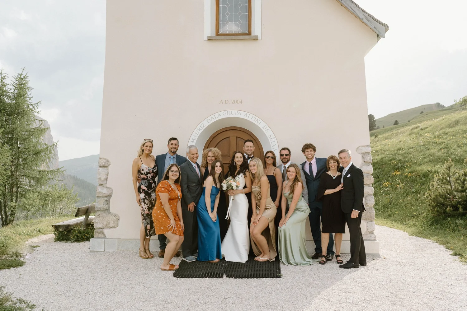 Group of people dressed in formal wedding attire standing in front of a small white chapel in a mountainous area