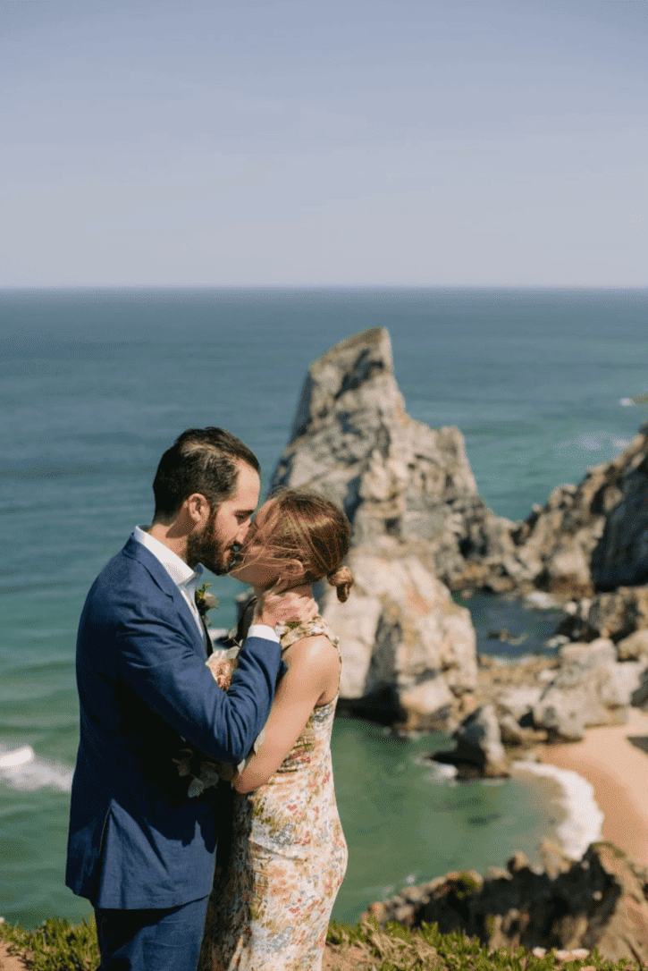 A couple dressed in wedding attire stands close to each other on a cliff by the ocean, with large rocks and a sandy beach in the background.