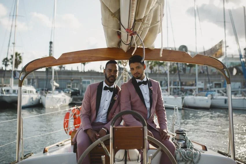 Two men in pink tuxedos sit on a boat at a marina, with sailboats and yachts in the background.