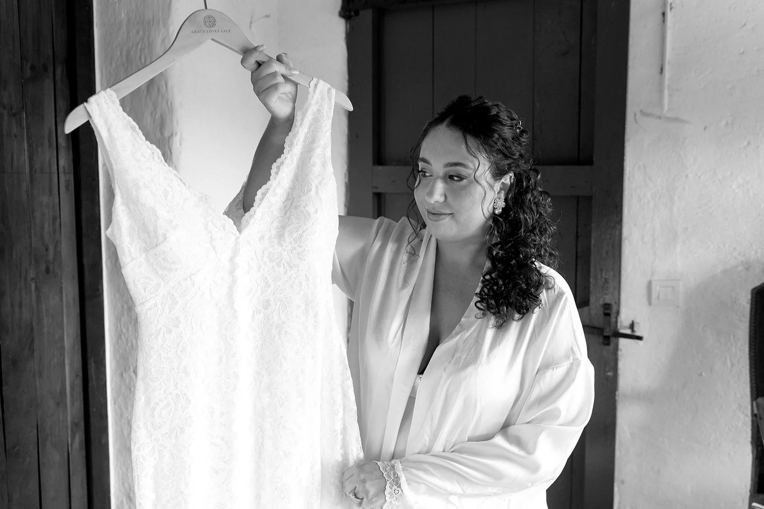 Women in a silk robe looking at a lace wedding dress on a hanger.