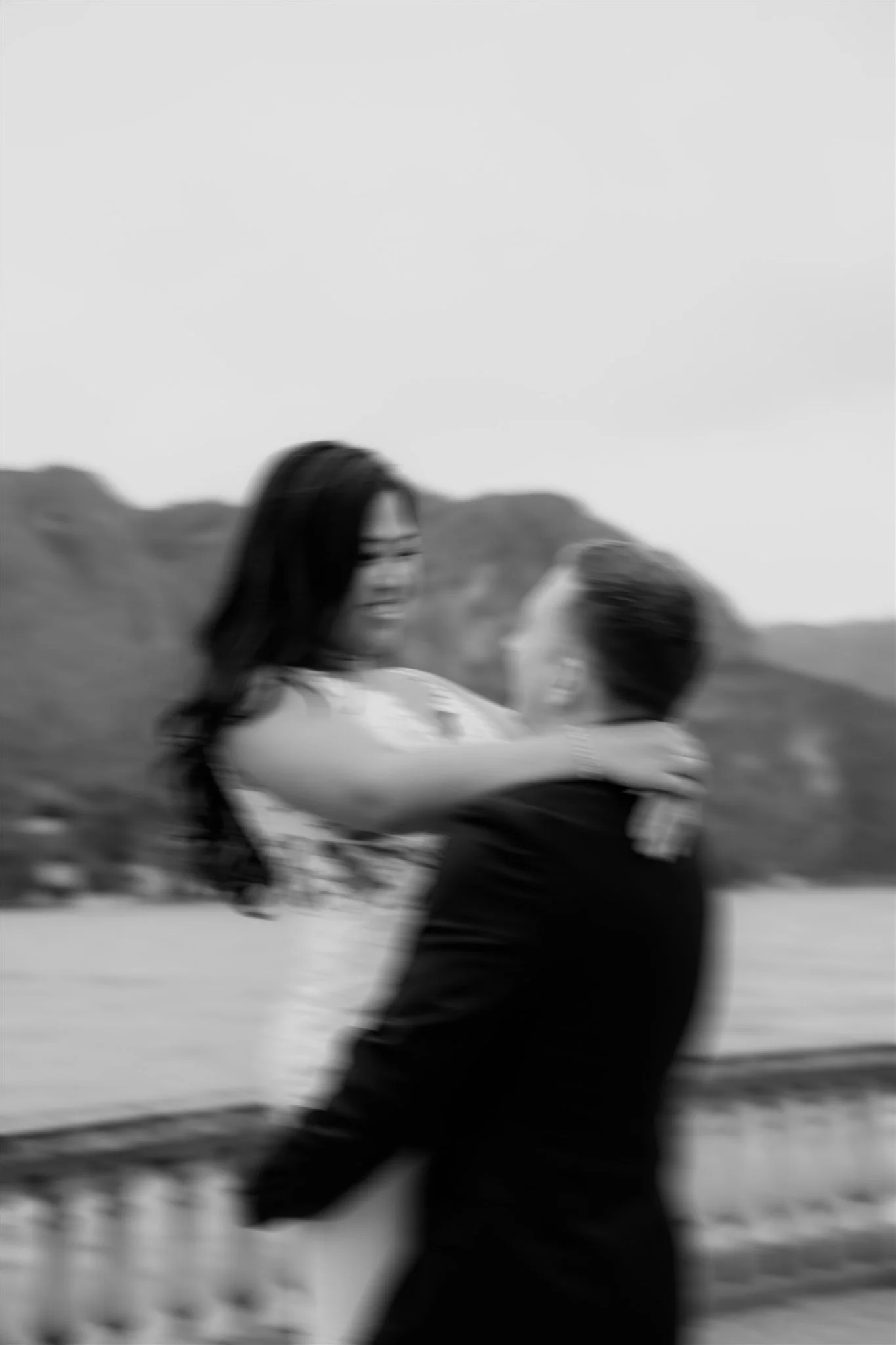 Black and white photo of two women hugging outdoors with mountains in the background.