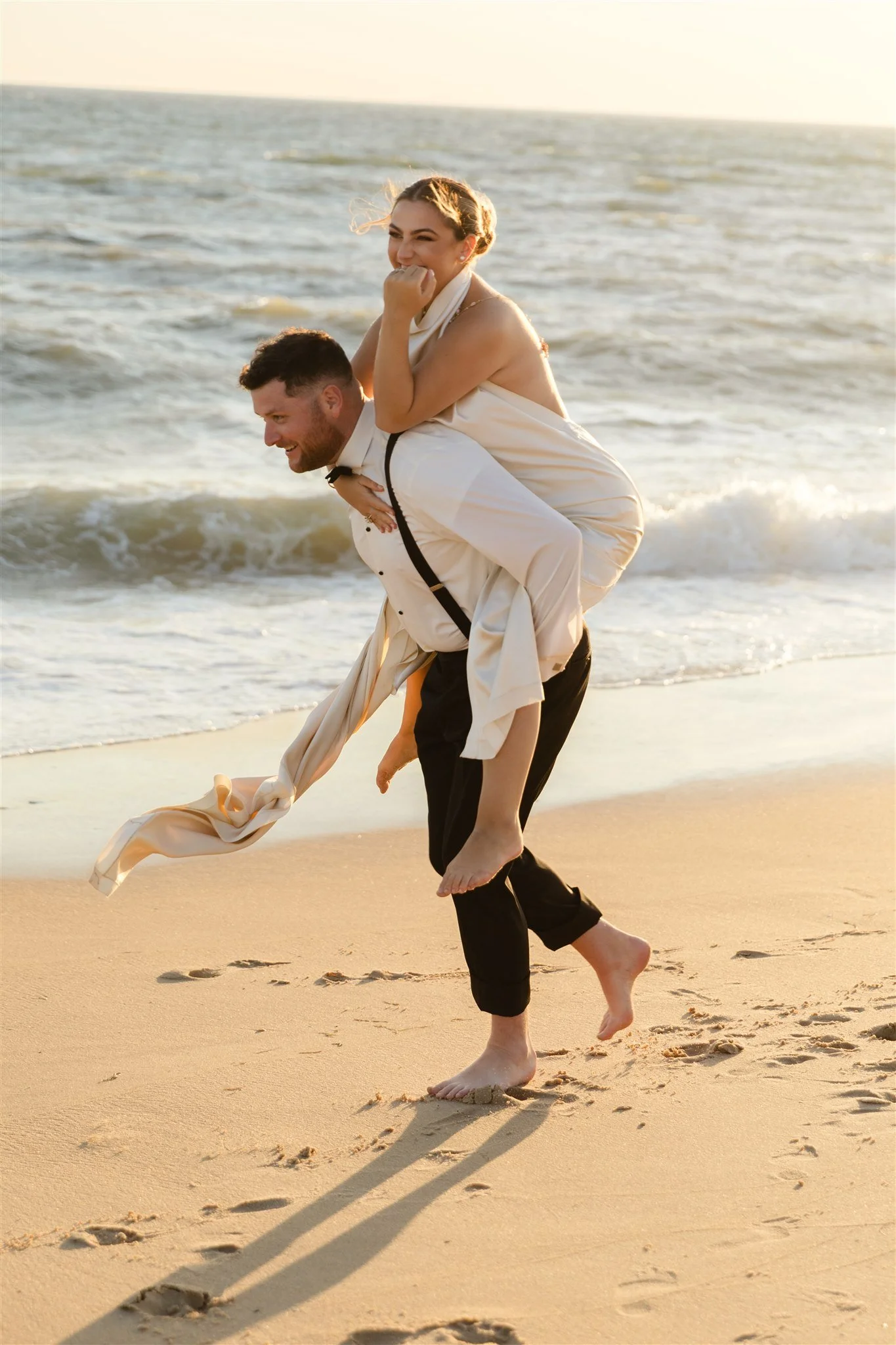 A man is giving a woman a piggyback ride on the beach during sunset. The woman is smiling and appears to be laughing, wearing a beige dress, while the man is dressed in formal black pants, a white shirt, and suspenders. They both are barefoot, and th