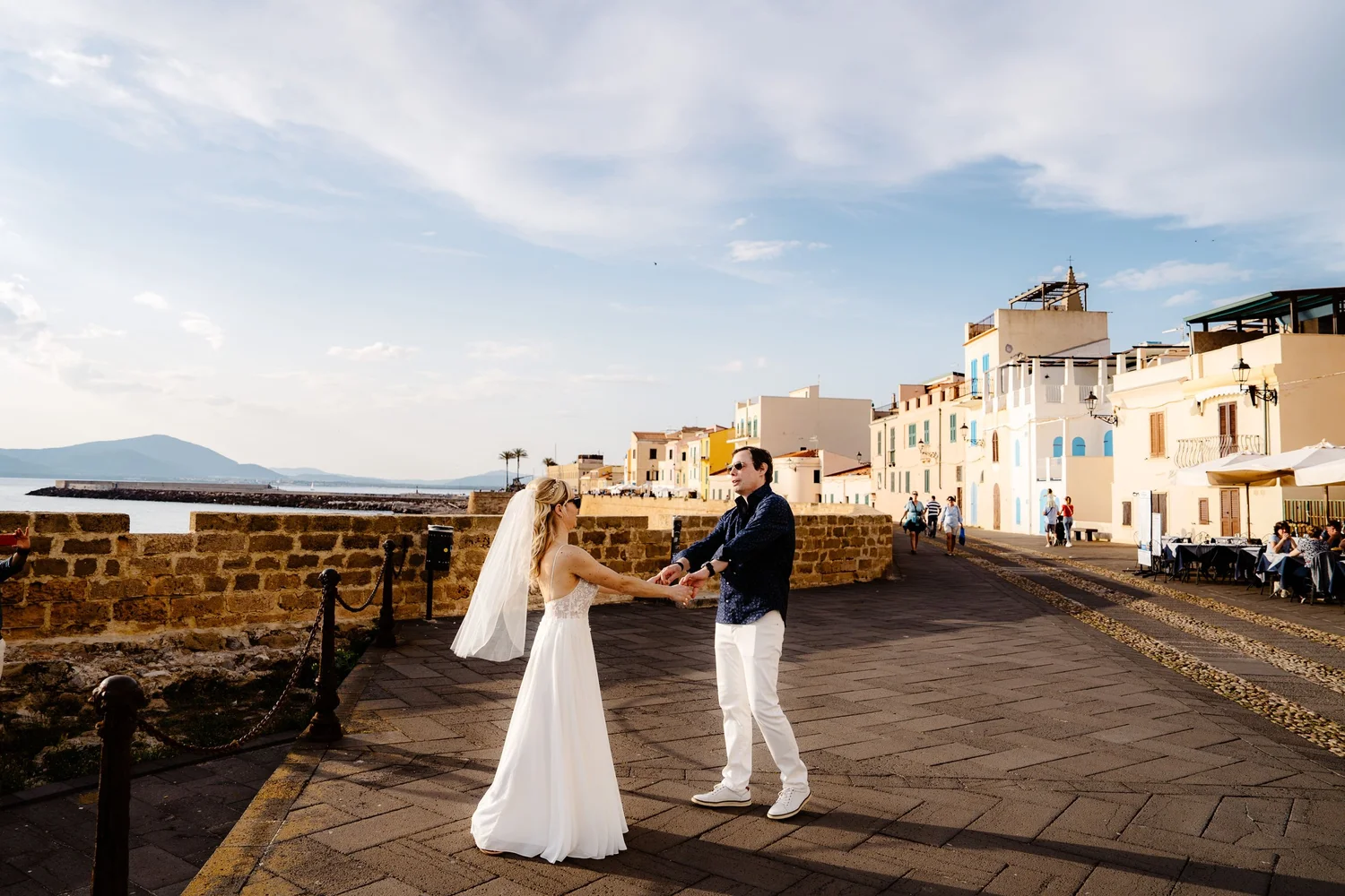 A bride and groom holding hands and dancing outdoors near a waterfront promenade, with colorful buildings and outdoor dining tables in the background, during late afternoon or early evening.