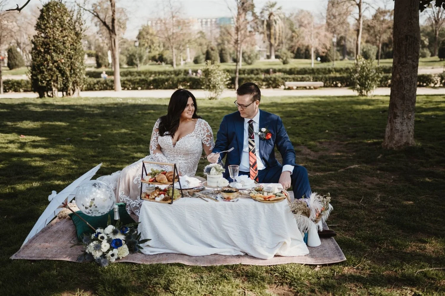 A couple dressed in wedding attire having a picnic in a park with food, drinks, and decorations on a blanket.