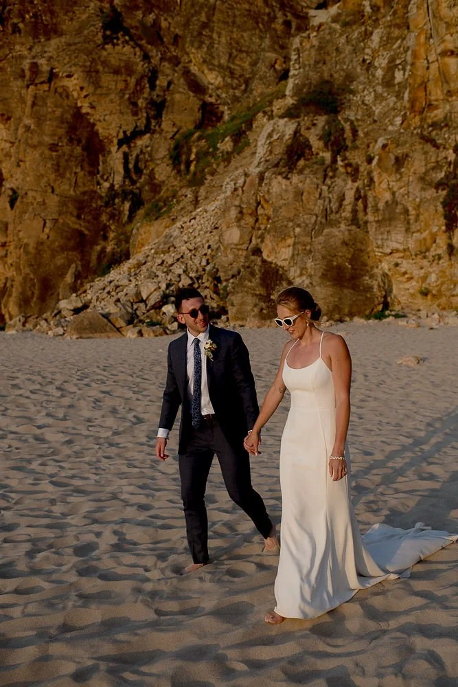 A couple dressed in wedding attire holding hands on a sandy beach with cliffs in the background, the man in a black suit and the woman in a white dress.