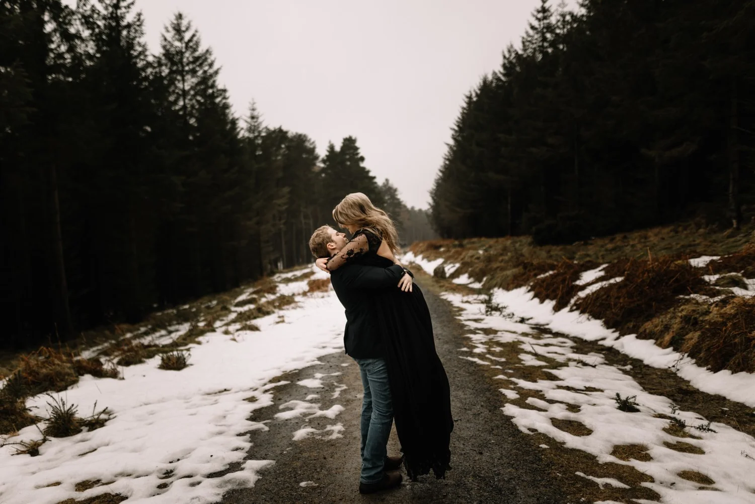 A couple hugging in a snowy forest road, with tall evergreen trees and overcast sky.