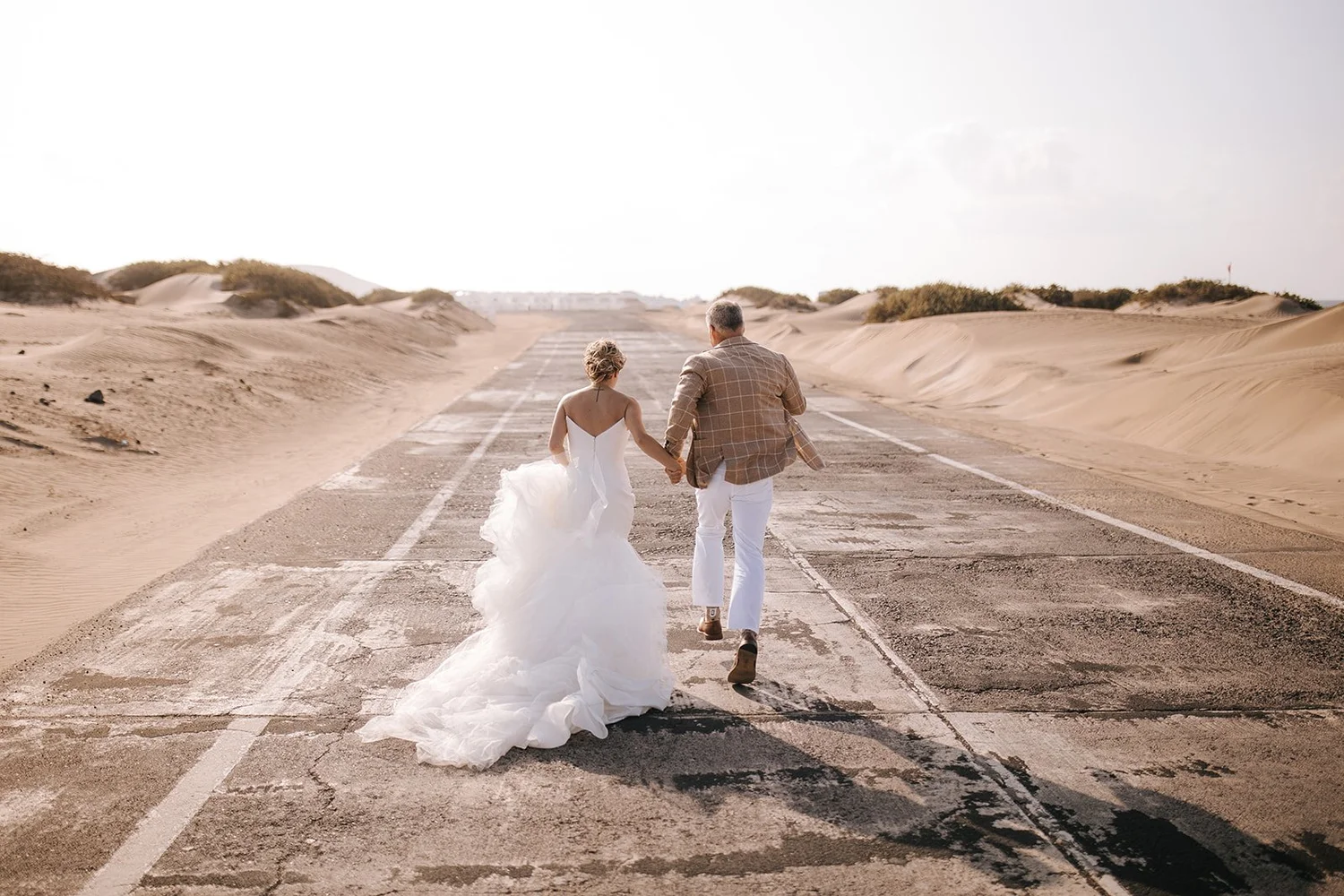 A couple in wedding attire walking hand in hand on an empty, weathered road through a desert landscape with sand dunes on each side.
