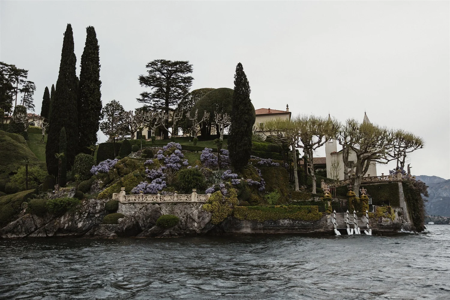 Lakeside garden with trimmed trees, flowering bushes, and a stone staircase, overlooking water with a rocky shoreline and mountainous background under a cloudy sky.