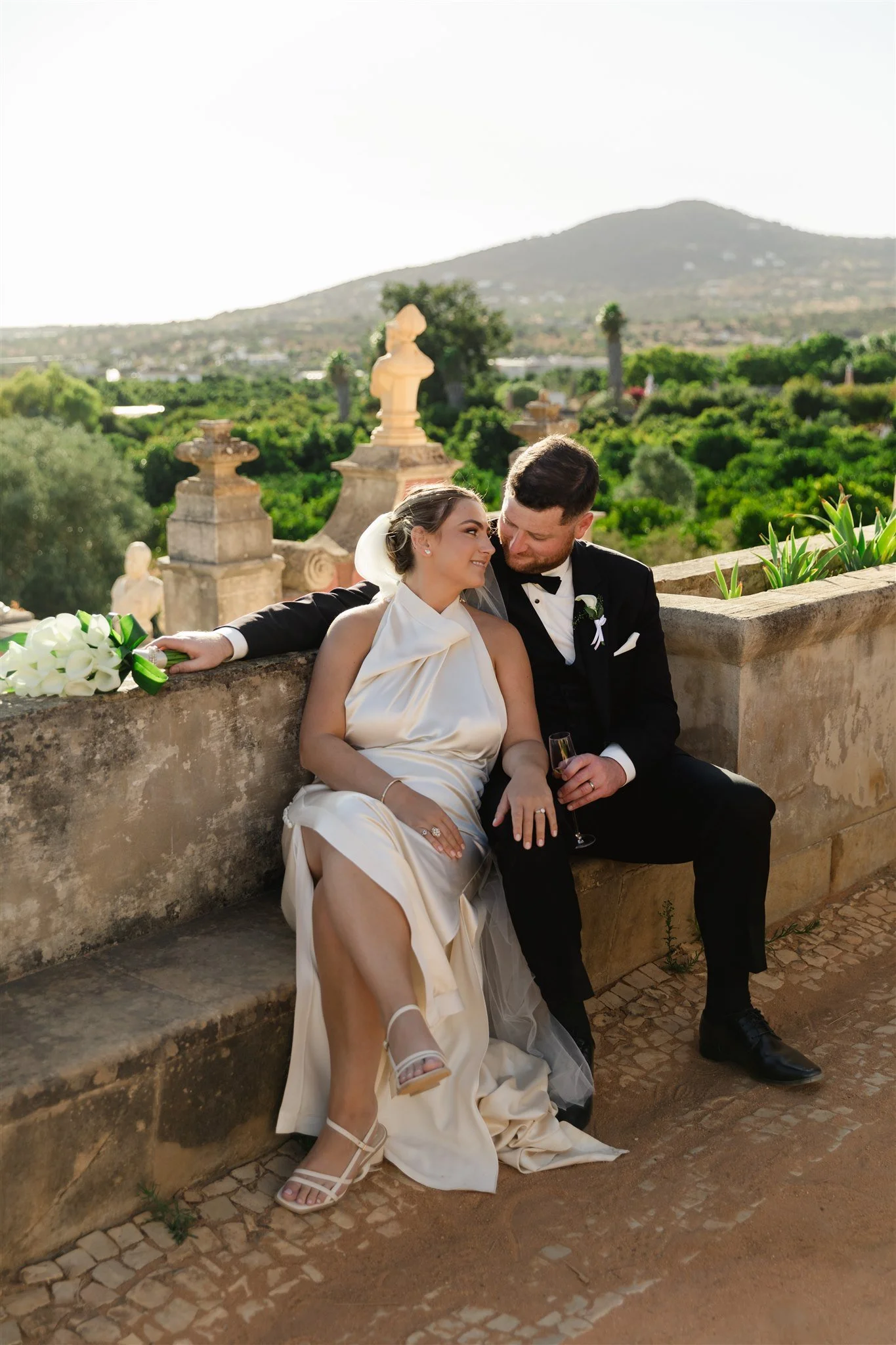 A bride and groom sitting on a stone wall with lush greenery and a mountain in the background. The bride wears a satin gown and white heels, holding a bouquet of calla lilies. The groom, in a tuxedo, holds a glass of champagne as they look at each ot