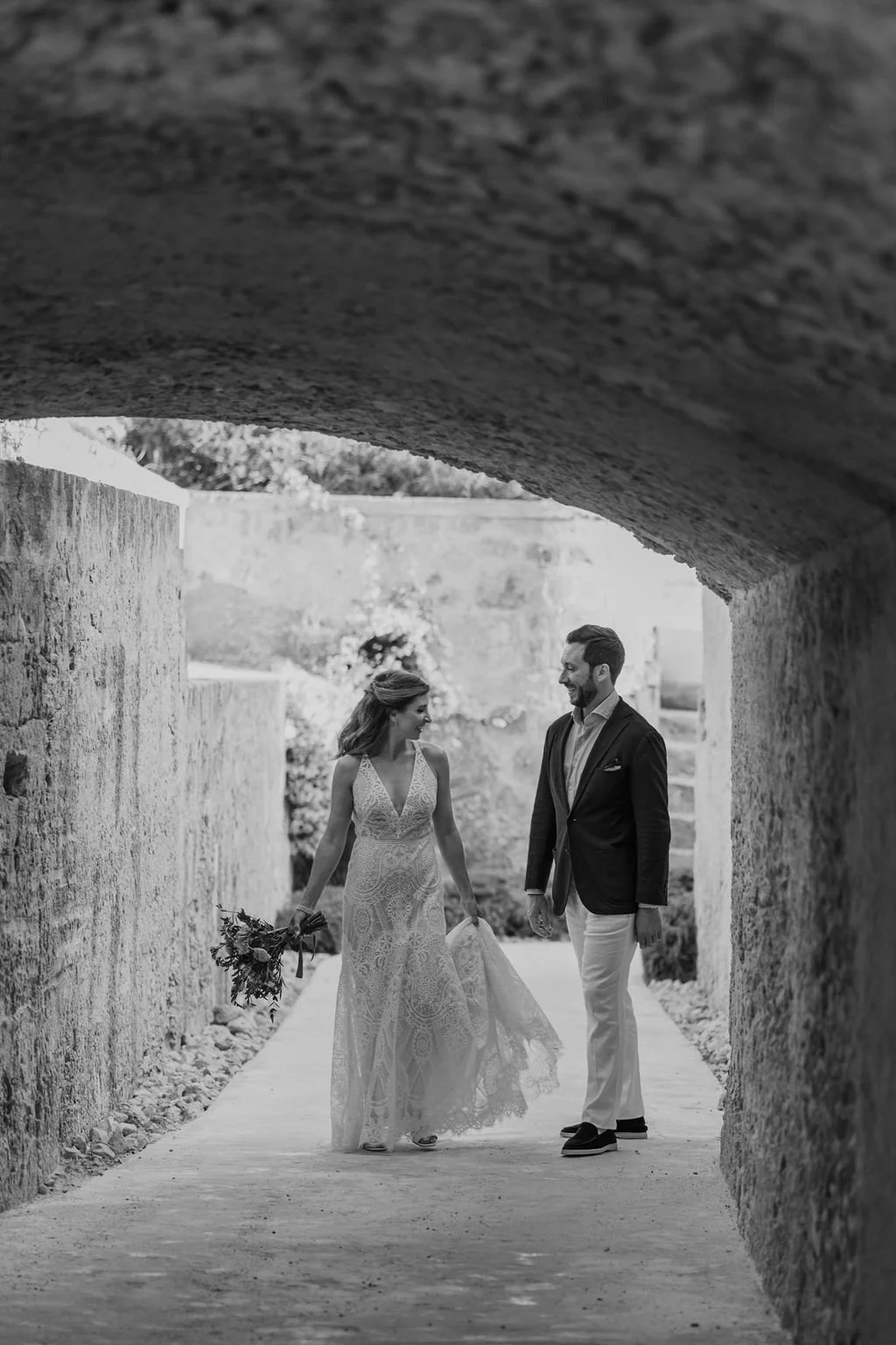 A bride and groom walking together in a tunnel, smiling at each other, with the bride holding a bouquet of flowers.