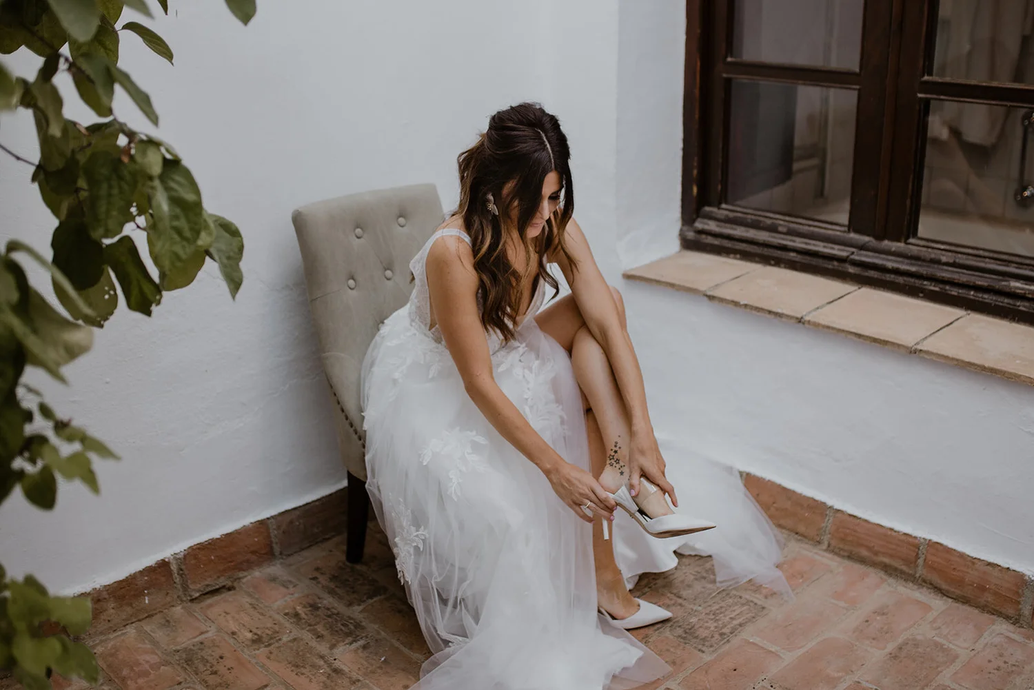 A woman in a wedding dress sits on a chair near a window, putting on high heels.