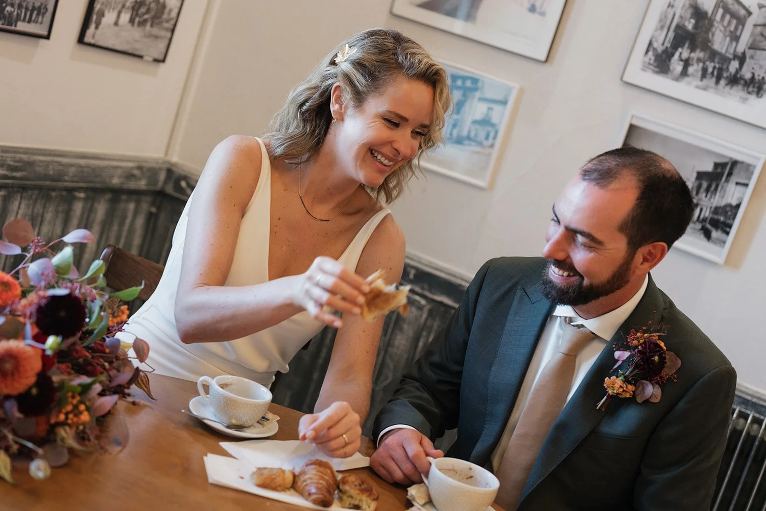 A woman and a man enjoying breakfast together at a restaurant. The woman is smiling and holding a piece of pastry, while the man is laughing. There are cups of coffee, pastries, and a flower arrangement on the table.