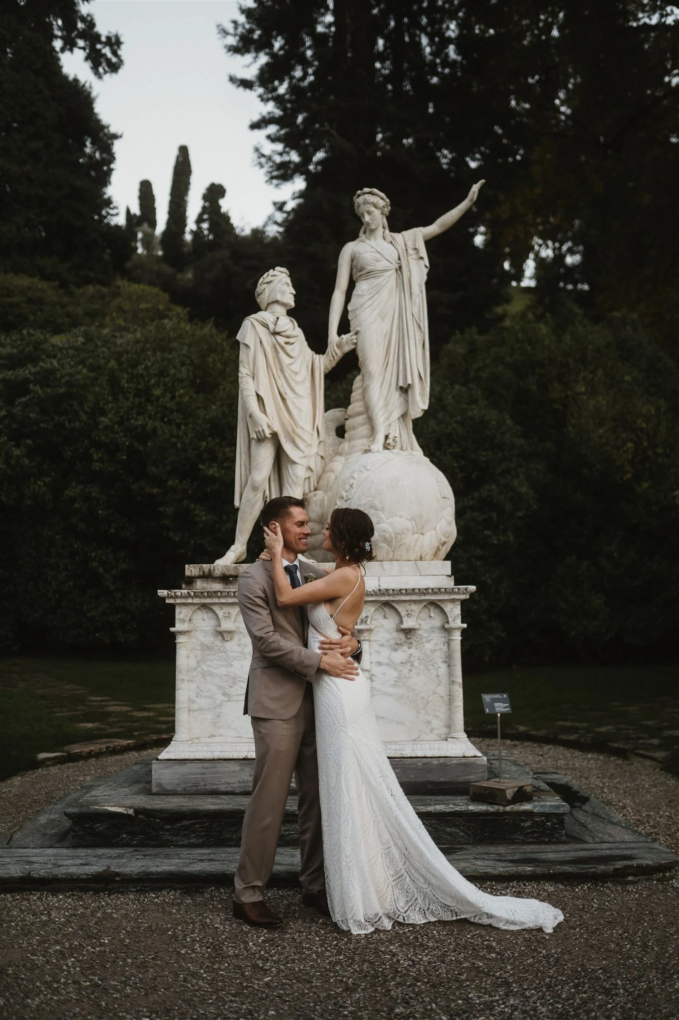 A bride and groom in wedding attire standing closely and looking into each other's eyes in front of a large marble statue of two women, one with a raised arm, in an outdoor garden setting during dusk.