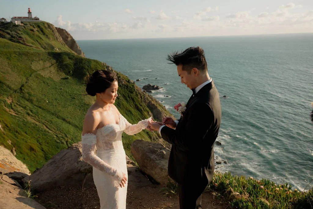 A bride and groom exchanging vows on a cliffside overlooking the ocean, with a lighthouse in the distance.