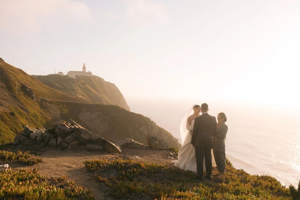 A wedding ceremony taking place on a cliffside with the ocean in the background and a lighthouse on a distant hill.