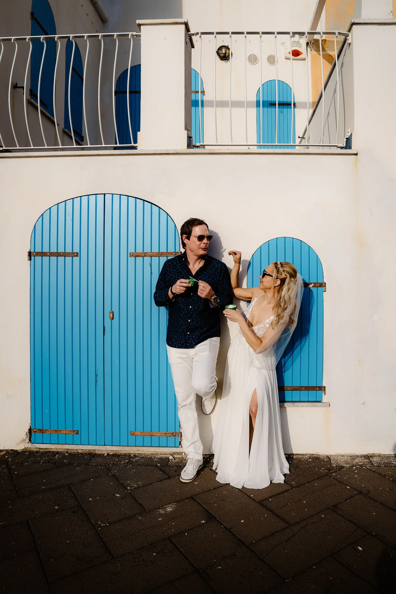A man and woman dressed in wedding attire standing in front of a white building with blue doors. The man is wearing sunglasses, a dark shirt, and white pants, and is holding a small green cup. The woman is wearing a bridal veil, sunglasses, and a whi