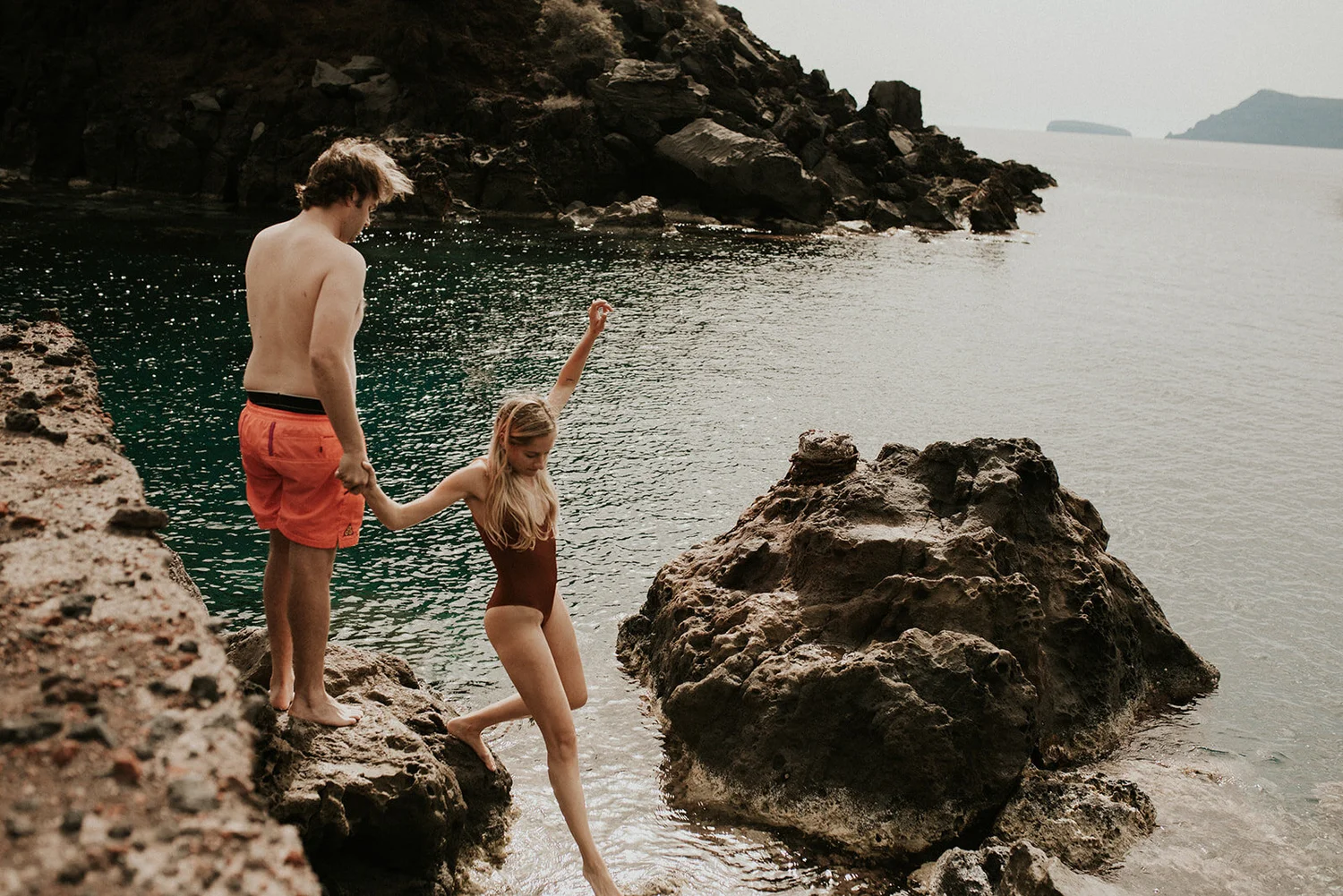 A man and a girl are stepping down from rocks into a body of water near a rocky shoreline, with distant landmasses on the horizon.