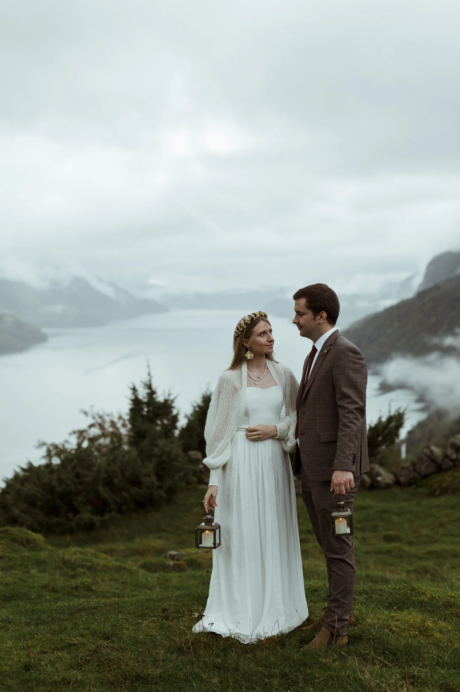 A bride and groom standing outdoors in a grassy area, holding lanterns, with mountains and a cloudy sky in the background.