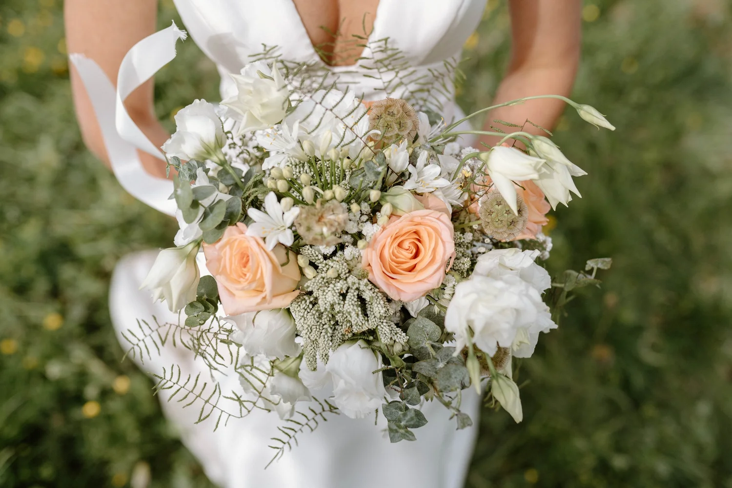 A person in a white dress holding a bouquet of white, peach, and cream-colored flowers with green foliage outdoors on green grass.