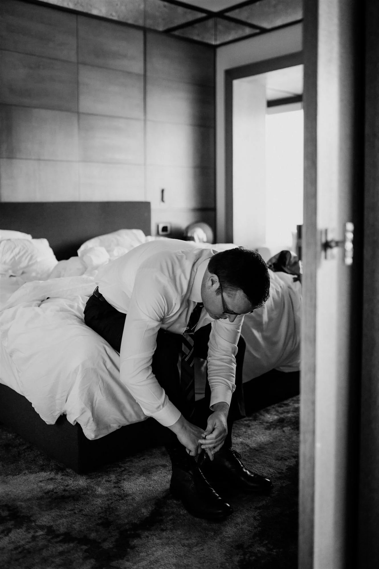 A man in a white shirt and tie is sitting on a bed in a hotel room, leaning forward and putting on black shoes. The room has a wooden headboard, and an open door shows bright daylight outside.