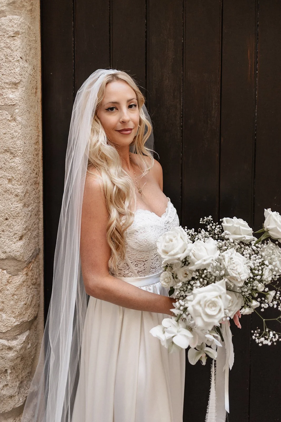 A bride with long blonde hair wearing a white strapless lace wedding dress and veil, holding a bouquet of white roses and baby's breath, standing in front of a dark wooden door and a stone wall.
